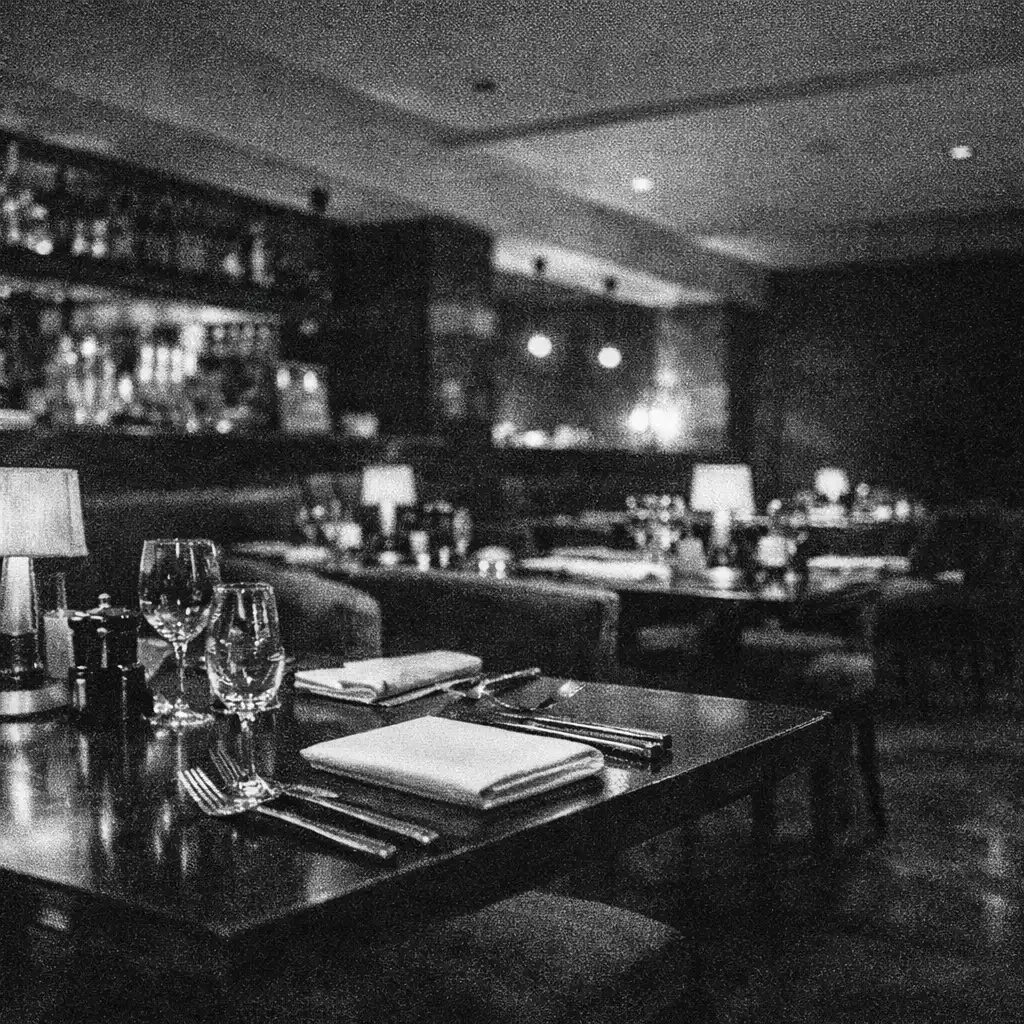 Wide view across prepared dining tables with folded napkins, silver cutlery and low lamps in a dark room.